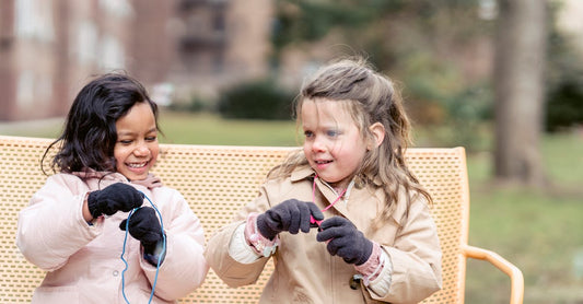 Cheerful little multiracial girlfriends in outerwear and gloves sitting together on bench and having fun with colorful earphones while spending spring day together in yard